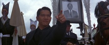 Movie still from “First Strike” (1996), directed by Stanley Tong – A man in a black suit points to a picture of an old man; Medium shot, Low angle