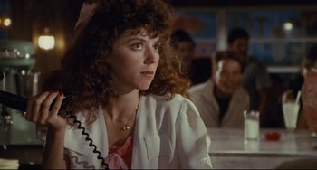 Movie still from “Porky's” (1981), directed by Bob Clark – A woman with curly hair is sitting in front of a group of people; Close Up shot, Over the shoulder angle