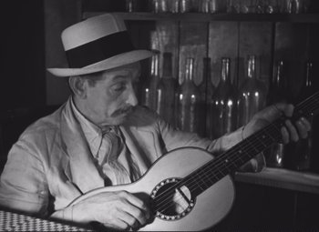 Movie still from “Port of Shadows” (1938), directed by Marcel Carné – An older man in a hat and a tie playing a guitar; Medium shot, Low angle