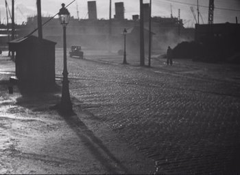 Movie still from “Port of Shadows” (1938), directed by Marcel Carné – A man walking down a street with a car in the background; Extreme Wide shot, High angle