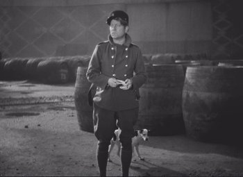Movie still from “Port of Shadows” (1938), directed by Marcel Carné – A man in a military uniform standing next to a dog; Wide shot, High angle