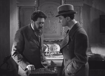 Movie still from “Port of Shadows” (1938), directed by Marcel Carné – Two men in suits and a hat are looking at each other in a room; Medium shot, High angle