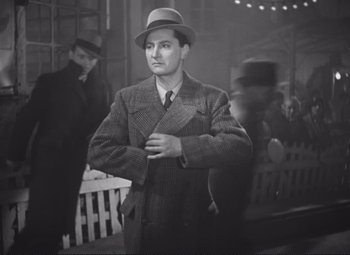Movie still from “Port of Shadows” (1938), directed by Marcel Carné – A man in a hat and a suit standing next to a fence; Medium shot, Low angle