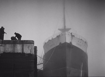 Movie still from “Port of Shadows” (1938), directed by Marcel Carné – A man sitting on top of a building next to a large ship; Extreme Wide shot, Low angle
