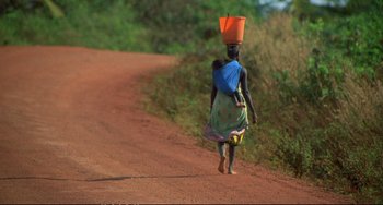 Movie still from “Powaqqatsi” (1988), directed by Godfrey Reggio – A woman walking down a dirt road carrying a bucket on top of her head; Wide shot, High angle