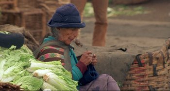 Movie still from “Powaqqatsi” (1988), directed by Godfrey Reggio – An older woman knitting on the side of the road; Close Up shot, High angle