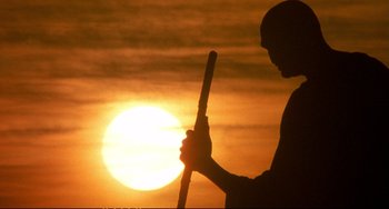 Movie still from “Powaqqatsi” (1988), directed by Godfrey Reggio – A man holding a baseball bat in front of a sunset; Medium shot, Low angle