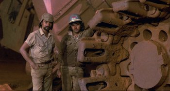 Movie still from “Powaqqatsi” (1988), directed by Godfrey Reggio – Two men standing in front of a pile of metal; Medium shot, High angle