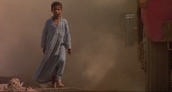 Movie still from “Powaqqatsi” (1988), directed by Godfrey Reggio – A young boy in pajamas standing in a dusty area; Wide shot, Low angle