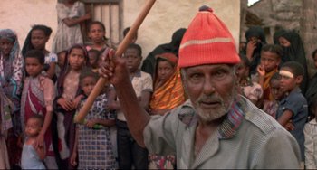 Movie still from “Powaqqatsi” (1988), directed by Godfrey Reggio – An old man wearing a red hat holding a wooden stick; Medium shot, Over the shoulder angle