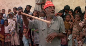 Movie still from “Powaqqatsi” (1988), directed by Godfrey Reggio – An old man holding a wooden stick in front of a group of people; Medium shot, Over the shoulder angle