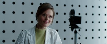 Movie still from “Predestination” (2014), directed by Michael Spierig – A woman wearing a lab coat smiles for the camera; Close Up shot, Over the shoulder angle