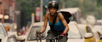 Movie still from “Premium Rush” (2012), directed by David Koepp – A woman riding a bike down a street; Medium shot, Low angle