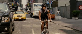 Movie still from “Premium Rush” (2012), directed by David Koepp – A man riding a bike down a street; Medium shot, Low angle