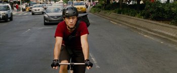 Movie still from “Premium Rush” (2012), directed by David Koepp – A man riding a bike down a street with cars in the background; Medium shot, Low angle