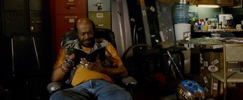 Movie still from “Premium Rush” (2012), directed by David Koepp – A man sitting in a chair while reading a book; Medium shot, High angle