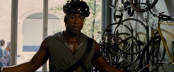 Movie still from “Premium Rush” (2012), directed by David Koepp – A man wearing a bicycle helmet in front of many bicycles; Medium shot, Over the shoulder angle