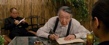 Movie still from “Premium Rush” (2012), directed by David Koepp – An older man sitting at a table writing on a notebook; Medium shot, Over the shoulder angle