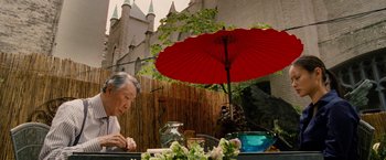 Movie still from “Premium Rush” (2012), directed by David Koepp – An older woman sitting at a table under an open red parasol; Medium shot, Over the shoulder angle