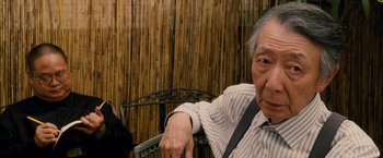 Movie still from “Premium Rush” (2012), directed by David Koepp – An older man sitting in a chair in front of a bamboo wall; Close Up shot, Over the shoulder angle