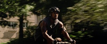 Movie still from “Premium Rush” (2012), directed by David Koepp – A man riding a bike on top of a lush green field; Medium shot, Low angle
