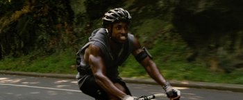 Movie still from “Premium Rush” (2012), directed by David Koepp – A man riding a bike down a street with trees in the background; Medium shot, Low angle
