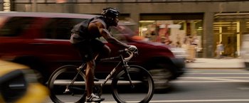 Movie still from “Premium Rush” (2012), directed by David Koepp – A man riding a bike down a street past a red car; Medium shot, Low angle