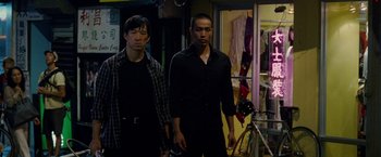 Movie still from “Premium Rush” (2012), directed by David Koepp – Two asian men standing next to each other in a room; Medium shot, Low angle