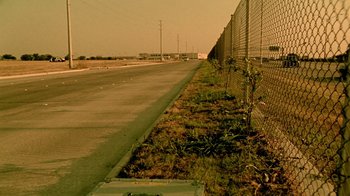 Movie still from “Primer” (2004), directed by Shane Carruth – A road with a fence and grass on the side of the road; Extreme Wide shot, Low angle