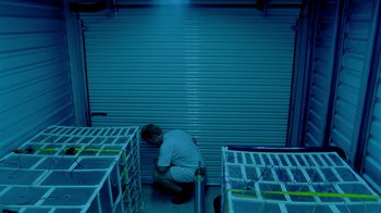 Movie still from “Primer” (2004), directed by Shane Carruth – A man kneeling down in front of a row of boxes; Wide shot, High angle