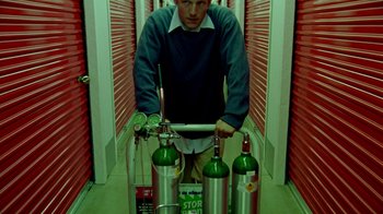 Movie still from “Primer” (2004), directed by Shane Carruth – A man holding a laptop in front of a row of wine bottles; Medium shot, Low angle