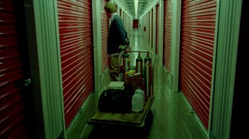 Movie still from “Primer” (2004), directed by Shane Carruth – A man pushing a cart full of luggage in a storage unit; Wide shot, High angle