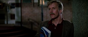 Movie still from “Prince of Darkness” (1987), directed by John Carpenter – A man holding a book while standing next to a brick wall; Close Up shot, Low angle