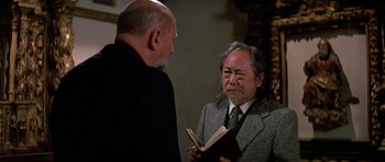 Movie still from “Prince of Darkness” (1987), directed by John Carpenter – Two men are talking to each other while one is holding a book; Medium shot, Over the shoulder angle