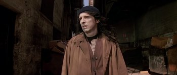 Movie still from “Prince of Darkness” (1987), directed by John Carpenter – A man with long black hair wearing a hat and a brown coat; Medium shot, Low angle