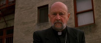 Movie still from “Prince of Darkness” (1987), directed by John Carpenter – An older man with a beard wearing a black suit; Close Up shot, Low angle