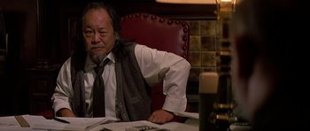 Movie still from “Prince of Darkness” (1987), directed by John Carpenter – A man sitting at a table with his arm resting on his shoulder; Medium shot, Low angle