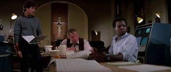 Movie still from “Prince of Darkness” (1987), directed by John Carpenter – Two men sitting at a table in front of a cross; Medium shot, Low angle