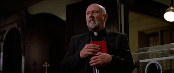 Movie still from “Prince of Darkness” (1987), directed by John Carpenter – A man in a black suit holding a red book; Close Up shot, Low angle