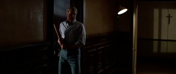 Movie still from “Prince of Darkness” (1987), directed by John Carpenter – A man standing in a dark room holding a wooden stick; Medium shot, Low angle