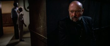 Movie still from “Prince of Darkness” (1987), directed by John Carpenter – An older man with a shaved head and a goatee is leaning against a wall; Close Up shot, Low angle