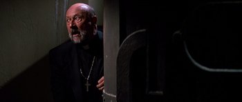 Movie still from “Prince of Darkness” (1987), directed by John Carpenter – An older man with a beard and a cross necklace; Close Up shot, Low angle