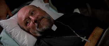 Movie still from “Prince of Darkness” (1987), directed by John Carpenter – An older man wearing a priest's outfit and a chain around his neck; Close Up shot, Low angle