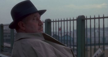 Movie still from “Prizzi's Honor” (1985), directed by John Huston – An older man wearing a hat sitting in front of a fence; Close Up shot, Over the shoulder angle