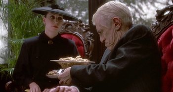 Movie still from “Prizzi's Honor” (1985), directed by John Huston – An older man holding a plate of food in front of an older woman; Medium shot, Over the shoulder angle