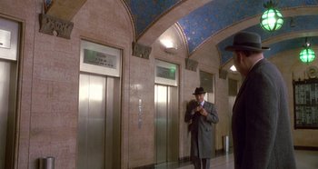 Movie still from “Prizzi's Honor” (1985), directed by John Huston – A man in a suit and a hat standing in front of two elevators; Wide shot, Low angle