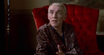 Movie still from “Prizzi's Honor” (1985), directed by John Huston – An older man sitting on a red chair talking; Medium shot, Over the shoulder angle