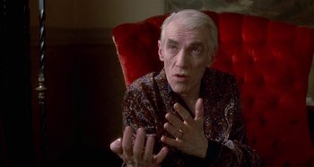 Movie still from “Prizzi's Honor” (1985), directed by John Huston – An older man sitting on a red chair with his hands up; Close Up shot, Over the shoulder angle
