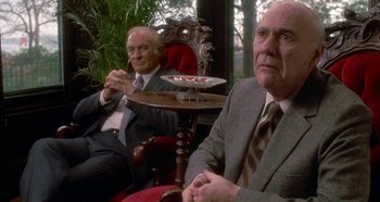 Movie still from “Prizzi's Honor” (1985), directed by John Huston – Two men in suits sitting at a table in front of a plant; Medium shot, Over the shoulder angle
