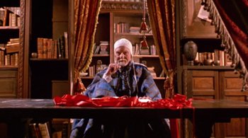 Movie still from “Prospero's Books” (1991), directed by Peter Greenaway – An old man sitting at a table in front of a red curtain; Medium shot, Low angle